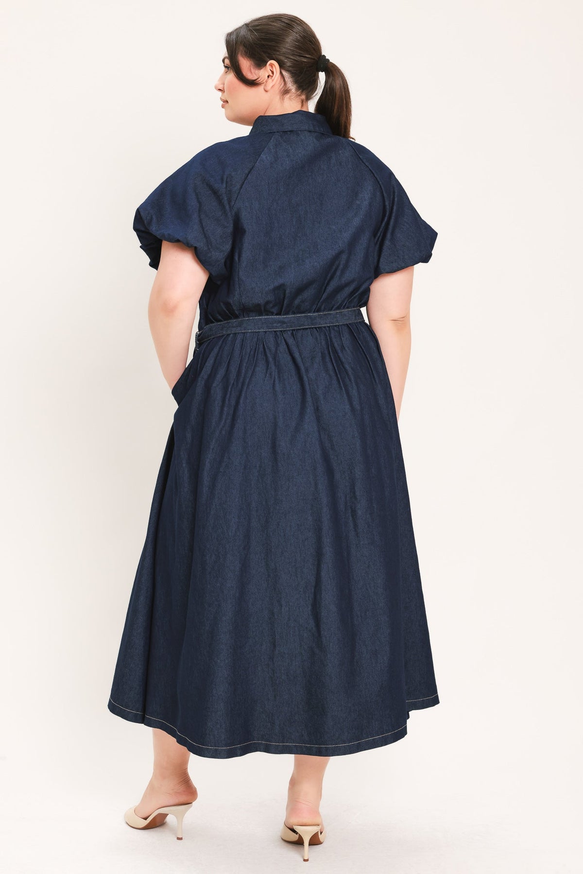 A woman wearing a dark navy blue midi dress with short puff sleeves and a matching fabric belt, shown from the back against a plain off-white studio background.
