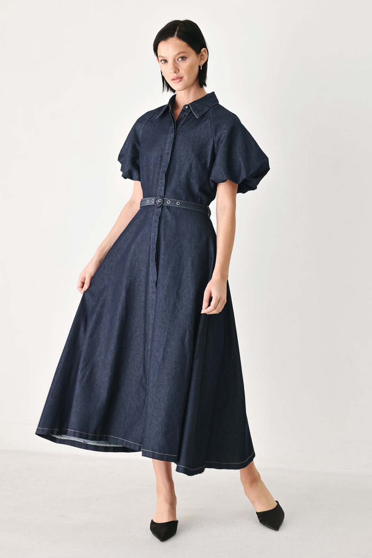 A woman modeling a dark navy blue midi shirt dress with puff sleeves, a pointed collar, and a matching fabric belt with silver-tone hardware, styled with black pointed-toe flats against a plain light gray studio background.