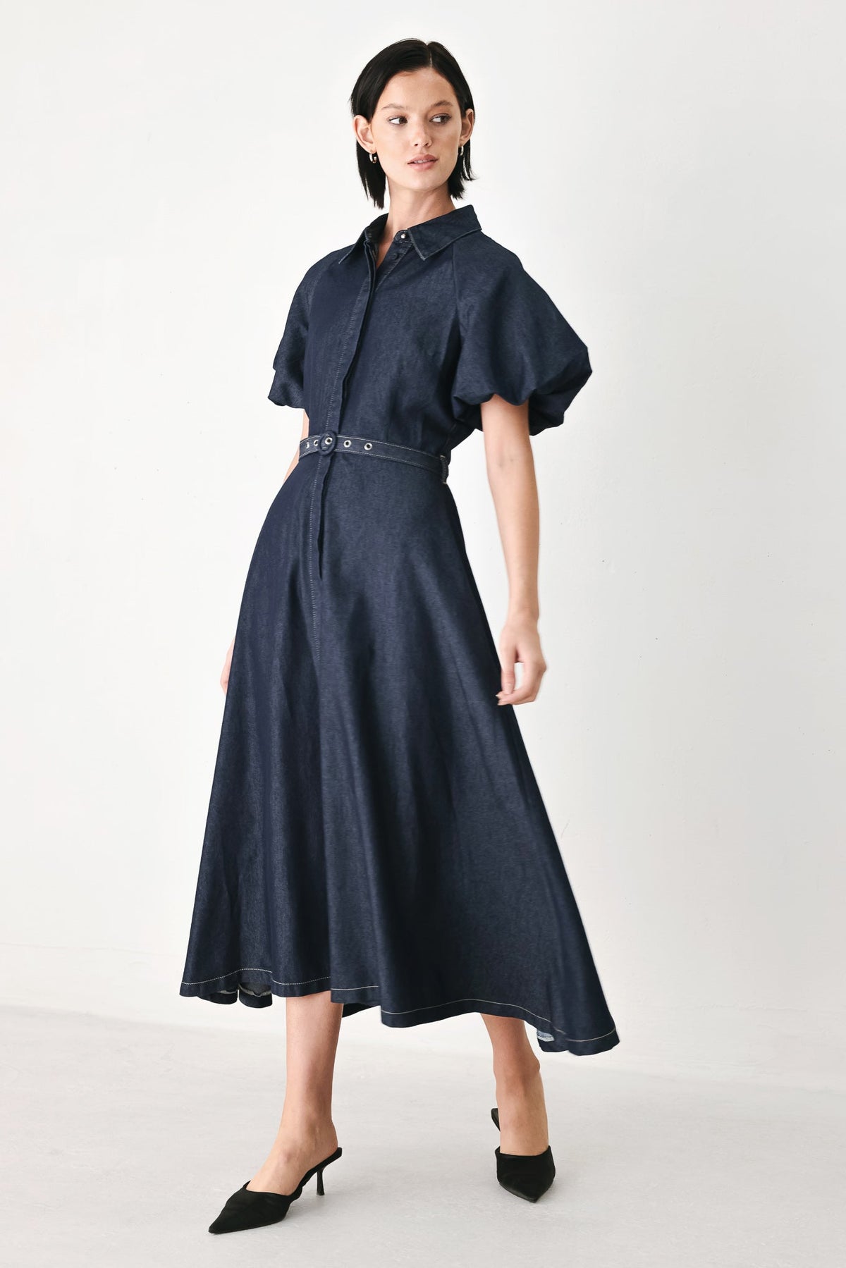 A model wearing a dark navy blue midi dress with short puff sleeves, a pointed collar, and a matching belt with silver studs, paired with black pointed-toe mule heels, set against a plain light gray studio background.