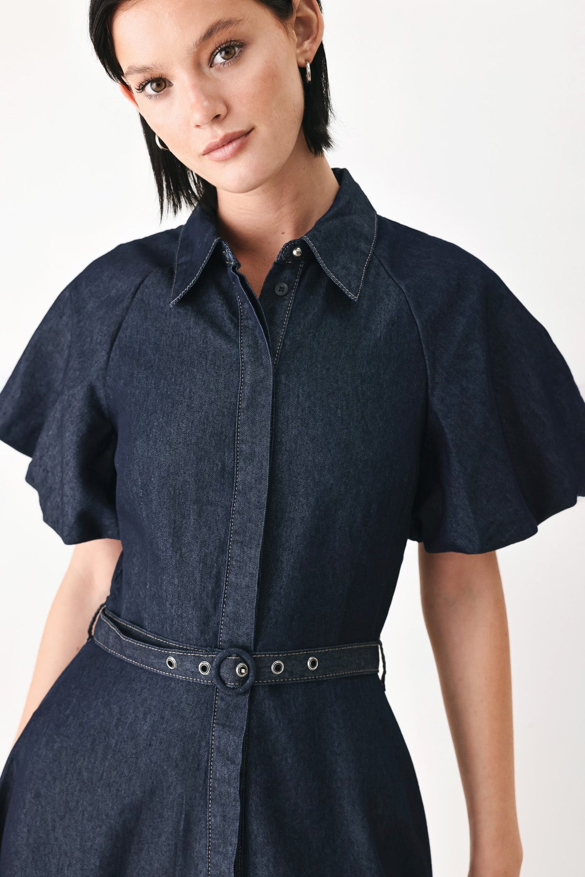 A woman wearing a dark indigo denim shirt dress with short flutter sleeves, a pointed collar, and a matching belt with silver-tone studs, photographed against a clean white studio background.