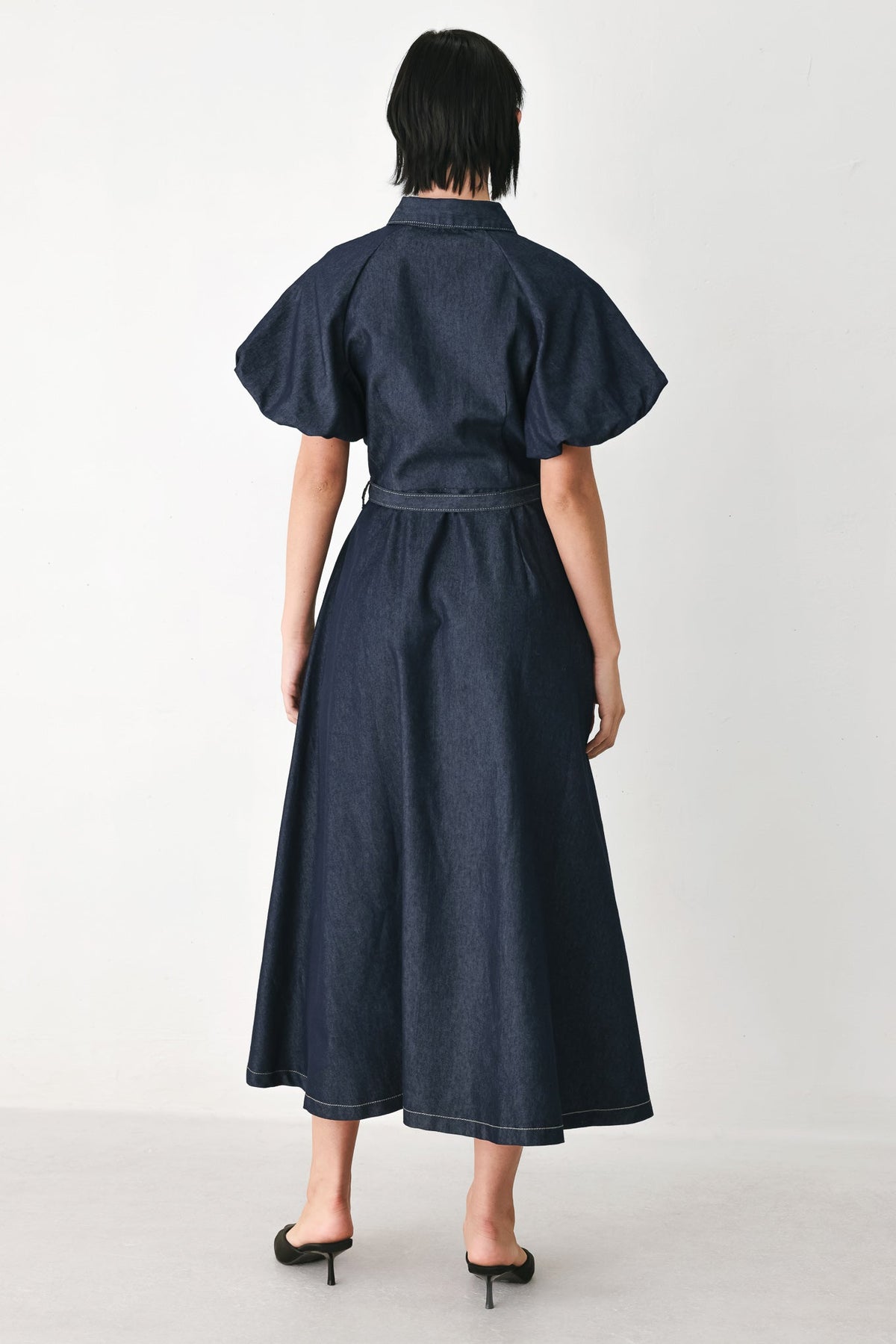 A model viewed from the back wearing a dark indigo denim midi dress with short puff sleeves, a high collar, and a matching belt, paired with black mule heels against a light gray studio background.