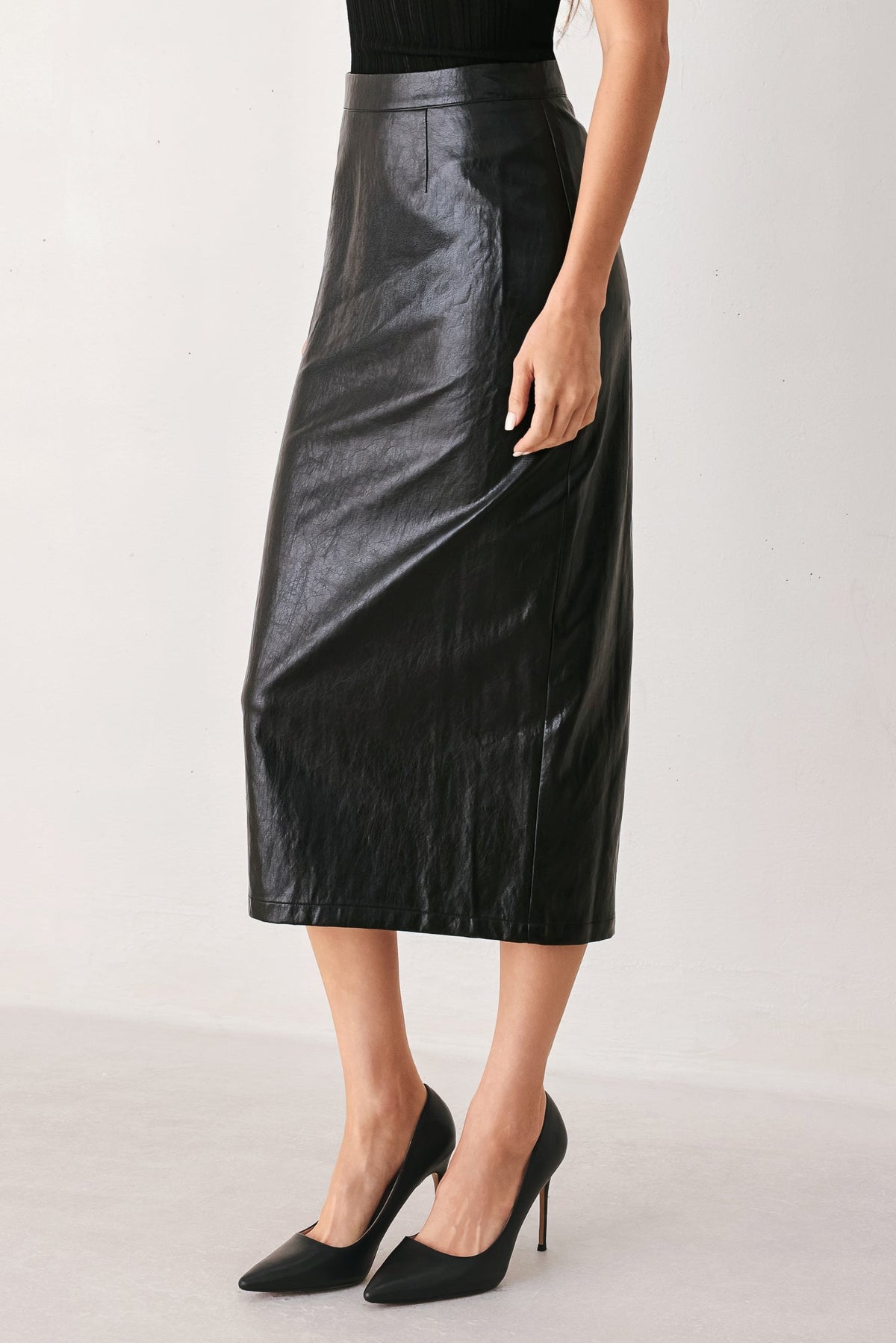 A model wearing a black faux-leather midi skirt with a subtle sheen, paired with black pointed-toe heels, shown from the waist down against a light gray studio background.