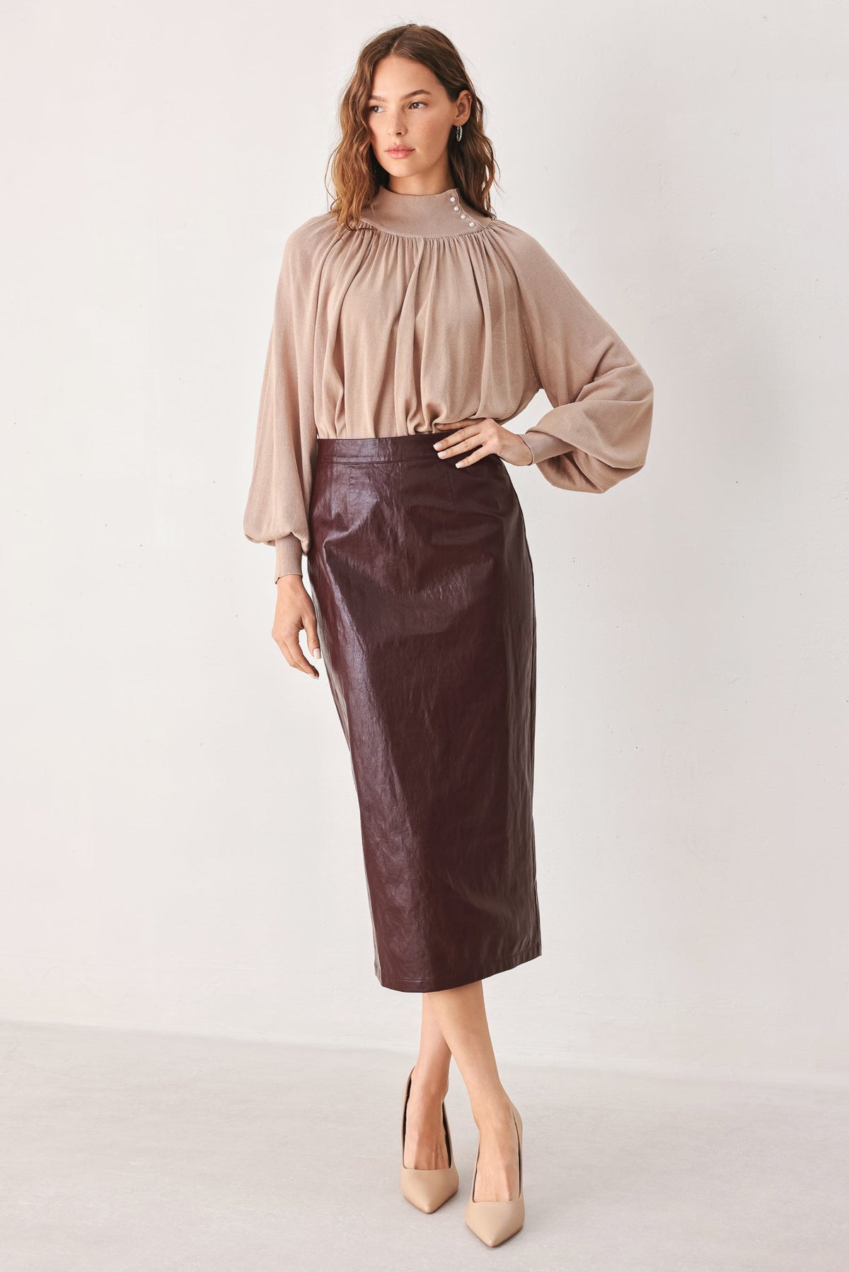 A woman modeling a beige long-sleeve pleated blouse and a dark brown faux-leather midi skirt, paired with beige heels, against a plain light gray studio background.