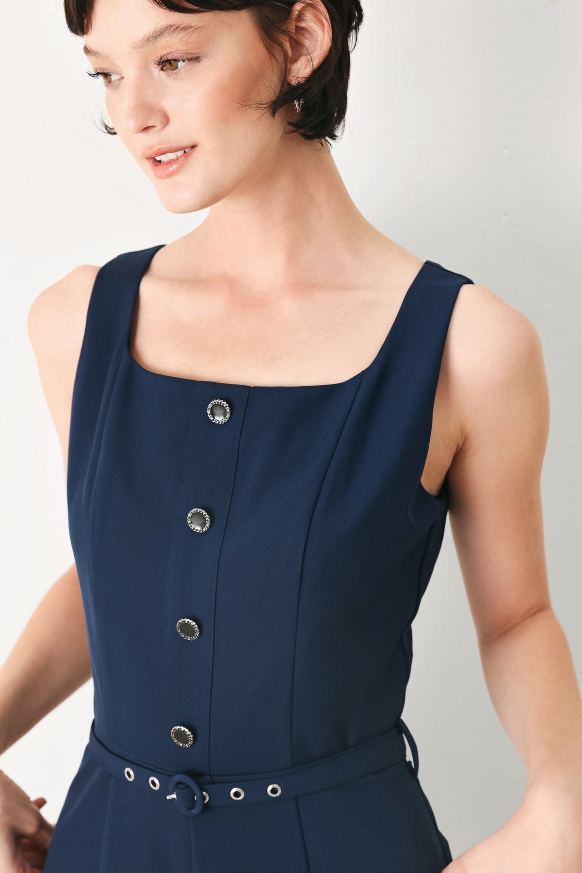 Woman wearing a navy blue sleeveless dress with button details on a plain background
