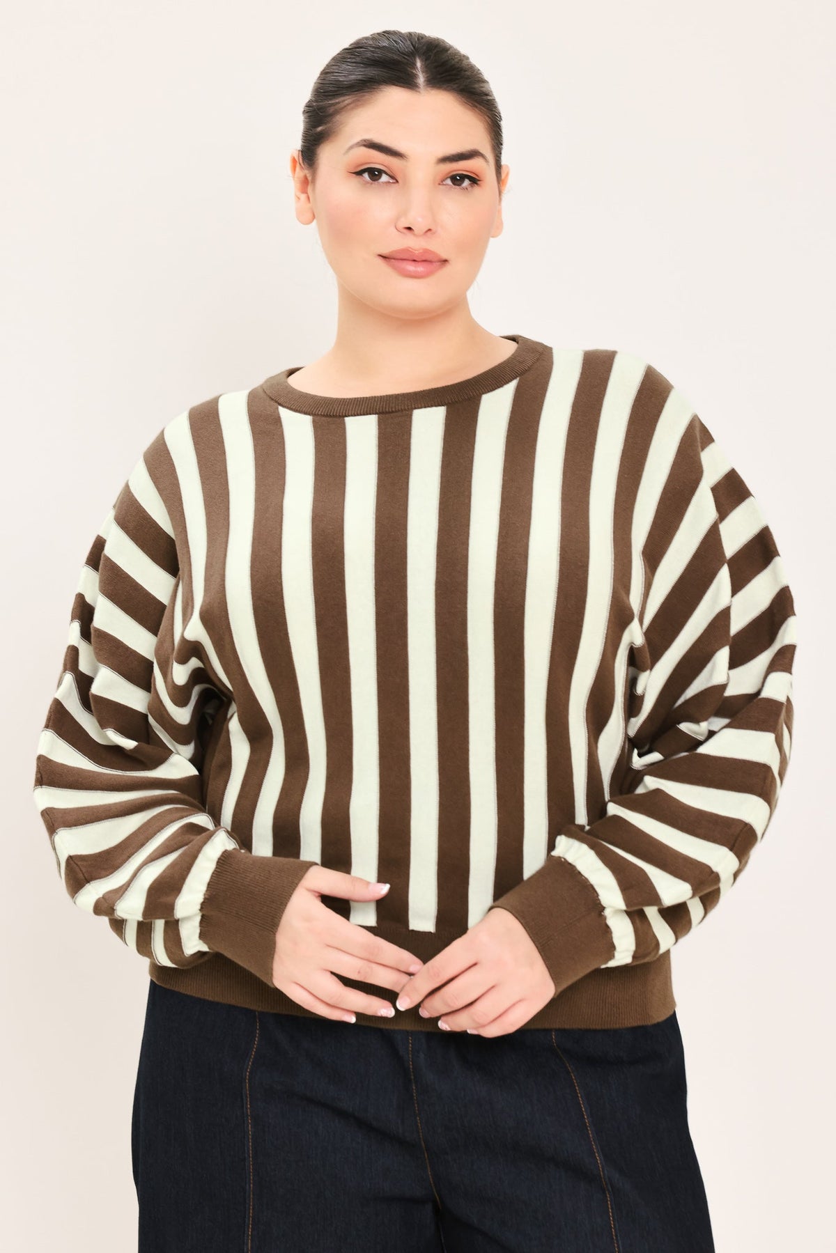 A woman wearing a brown and cream vertical striped knit sweater with long balloon sleeves and ribbed cuffs, paired with dark jeans, against a plain light background.