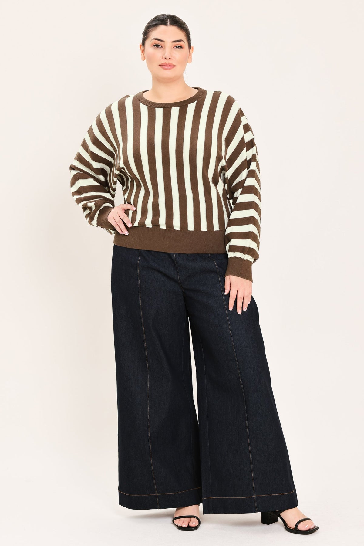 A woman modeling a brown and cream striped knit sweater with wide-leg dark denim pants and black sandals, standing against a plain light beige studio background.