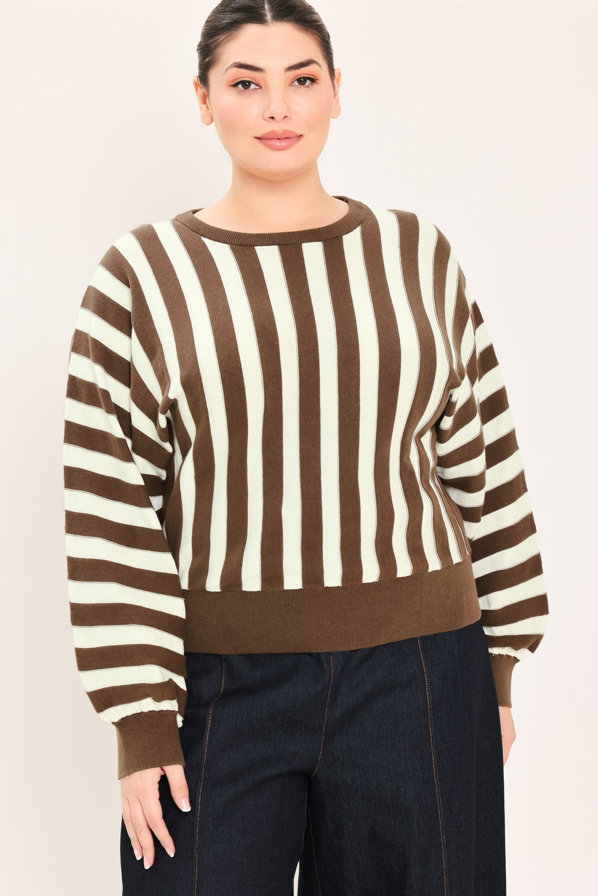 A woman wearing a brown and cream striped knit sweater with dark blue jeans, standing against a plain light beige studio background.
