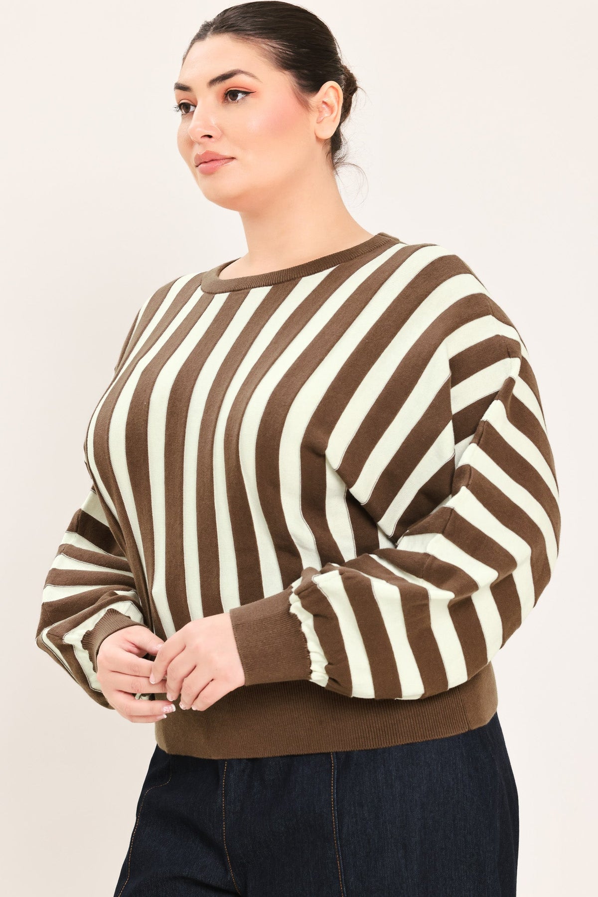 A woman wearing a brown and cream striped long-sleeve knit sweater, paired with dark blue jeans, against a plain light beige background.