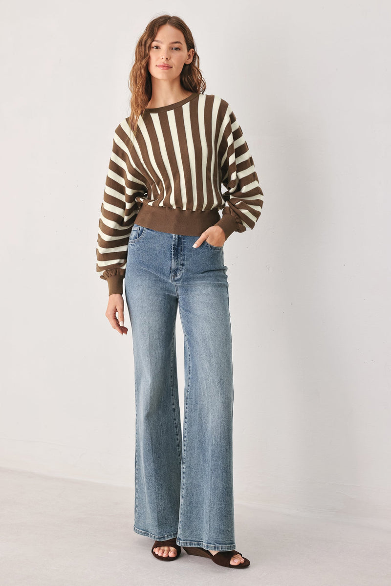 A woman modeling a brown and cream striped cropped sweater with long sleeves, paired with high-waisted, wide-leg blue jeans and dark brown slide sandals, against a plain light gray studio background.