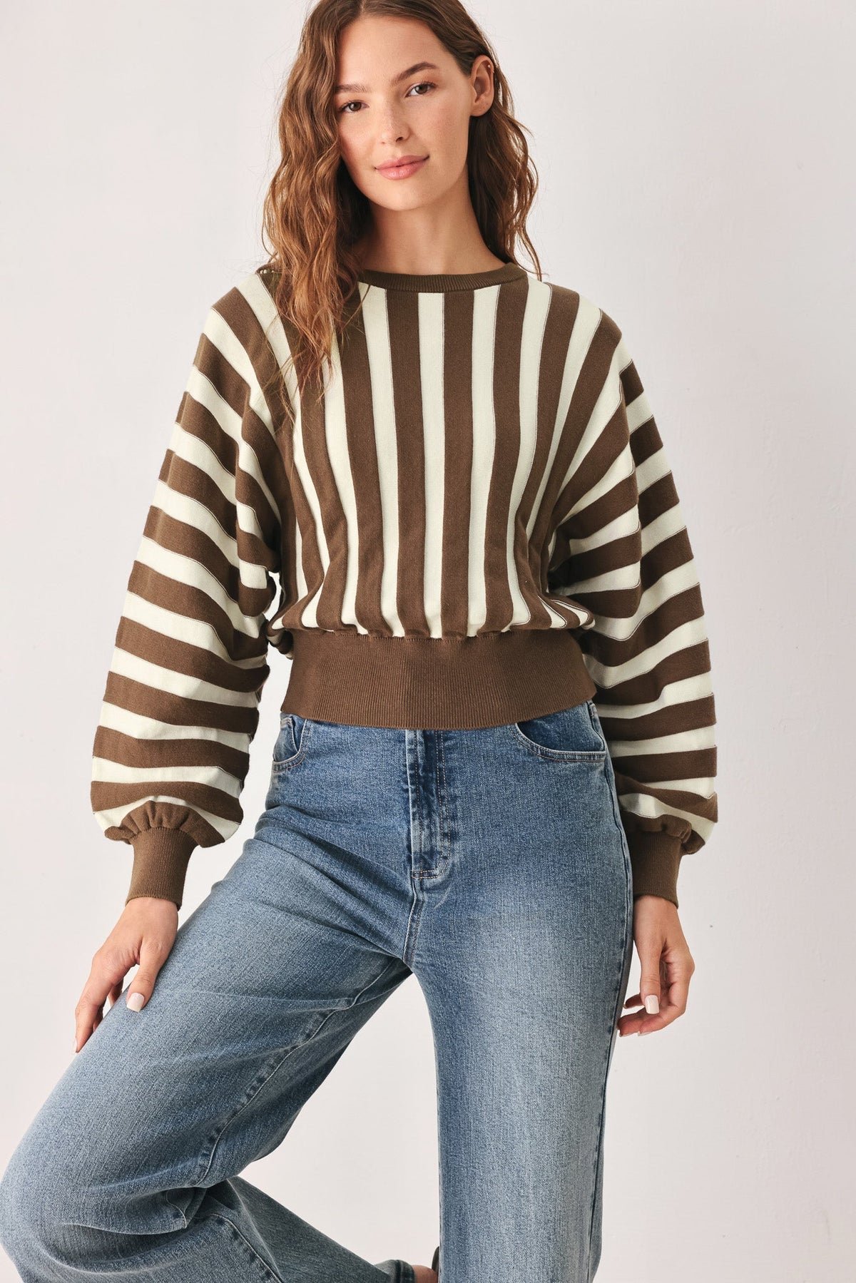A woman wearing a brown and cream striped long-sleeve knit sweater with balloon sleeves and a ribbed hem, paired with blue jeans, against a light gray studio background.