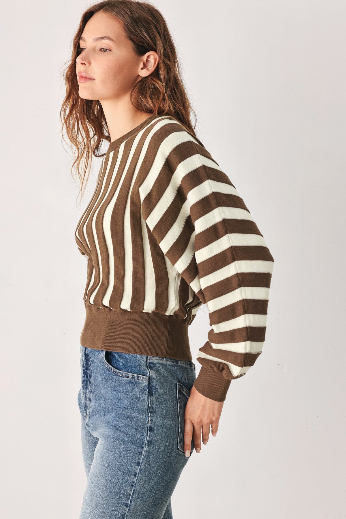 A woman wearing a brown and cream striped long-sleeve knit top with a ribbed waistband and cuffs, paired with blue jeans, photographed against a plain light gray studio background.