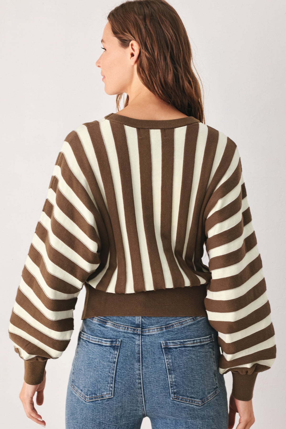 A woman shown from the back wearing a brown and cream striped cropped sweater with balloon sleeves, paired with blue jeans, against a light gray studio background.