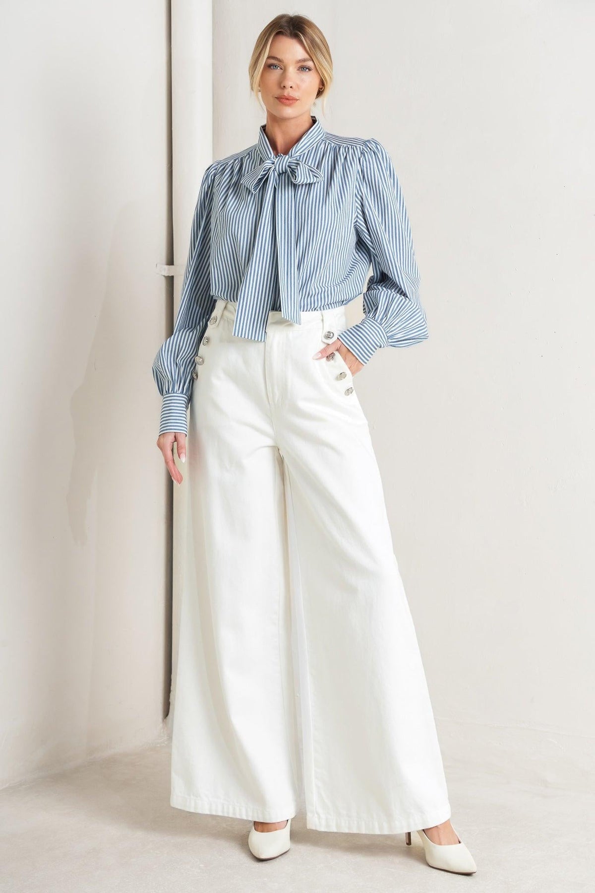 Woman wearing a blue striped blouse and white pants against a plain background