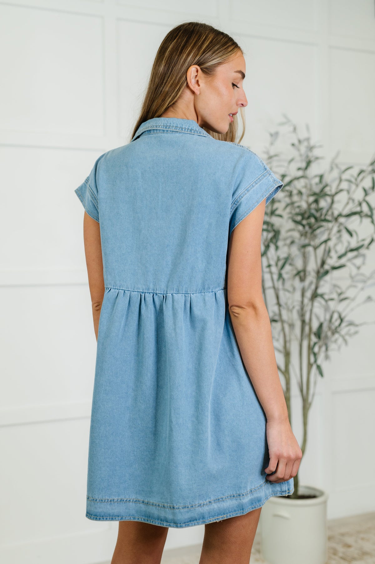 Woman wearing a light blue denim dress standing indoors with a white wall and plant in the background.