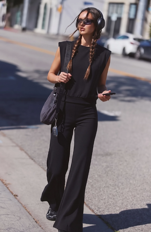 A woman walking on a sunny city street wearing a black sleeveless top and matching black wide-leg pants, with black headphones, sunglasses, and a dark shoulder bag.
