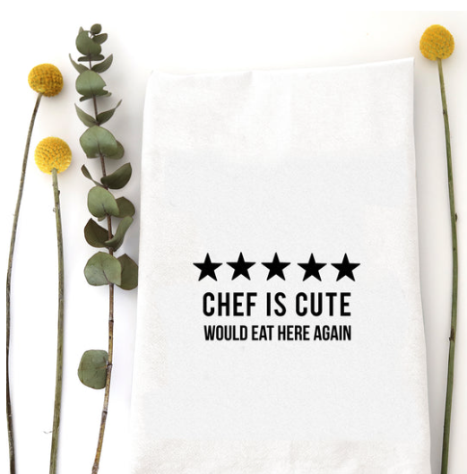 A white kitchen towel with black printed text reading 'CHEF IS CUTE WOULD EAT HERE AGAIN' beneath five stars, styled flat-lay with eucalyptus and yellow flowers on a white background.