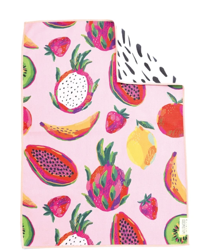 A square pink kitchen towel featuring a colorful fruit print, with a folded corner showing a white reverse side with black spots. The fruits include strawberries, dragon fruit, papayas, bananas, and lemons.