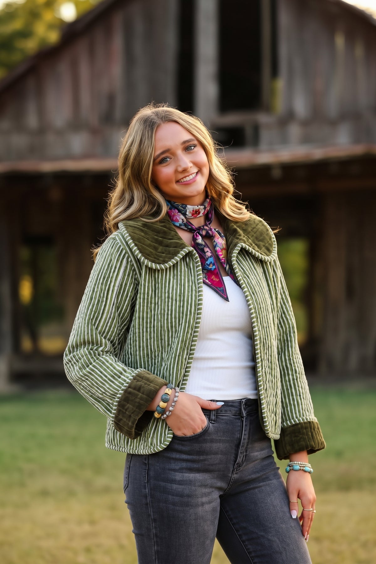 A woman stands outdoors in front of a rustic wooden barn, wearing a green corduroy cropped jacket with a dark green collar and cuffs, a white top, dark gray jeans, and a colorful floral neck scarf.