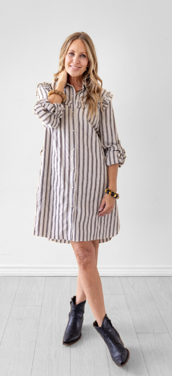 A woman modeling a beige and brown vertically striped button-down shirt dress with ruffle details on the shoulders, paired with dark blue leather ankle boots, standing in a bright room with a white wall and light wood floor.