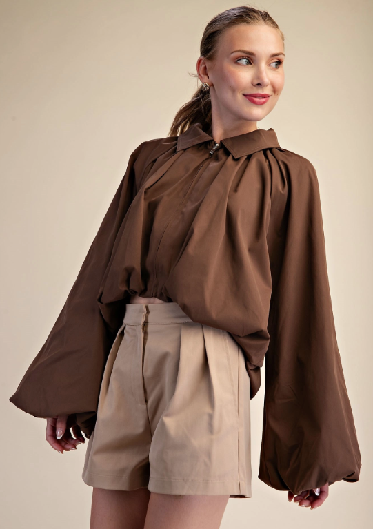 A woman modeling a brown long-sleeve blouse with dramatic balloon sleeves and a gathered neckline, paired with beige shorts, against a plain beige studio background.