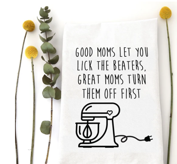 A white kitchen towel with black printed text and a mixer illustration, styled flat-lay with eucalyptus stems and yellow pom-pom flowers on a white background.