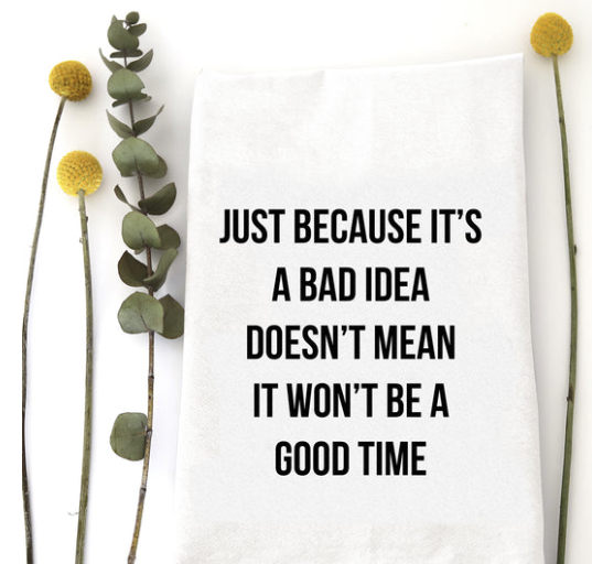 A white fabric tea towel with black printed text reading 'JUST BECAUSE IT'S A BAD IDEA DOESN'T MEAN IT WON'T BE A GOOD TIME', styled flat-lay with eucalyptus stems and yellow flowers on a white background.