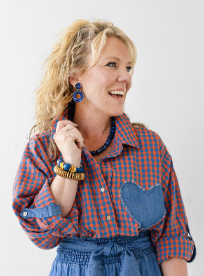 A woman with blonde curly hair wearing a red and blue gingham button-up shirt with a denim heart patch pocket, blue denim shorts, and colorful beaded jewelry, posing against a light gray studio background.