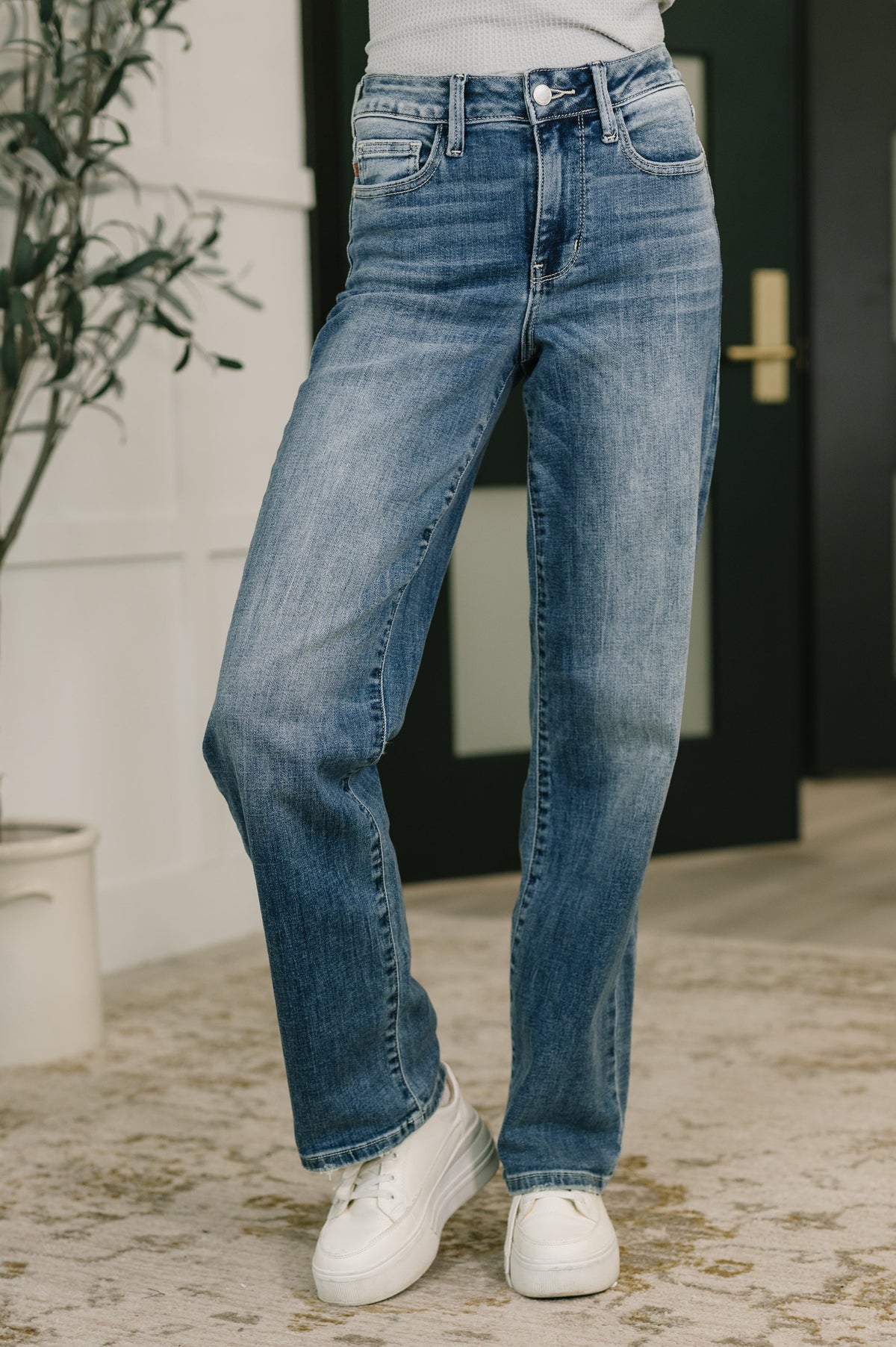 Person wearing blue jeans and white sneakers standing indoors with a plant in the background.