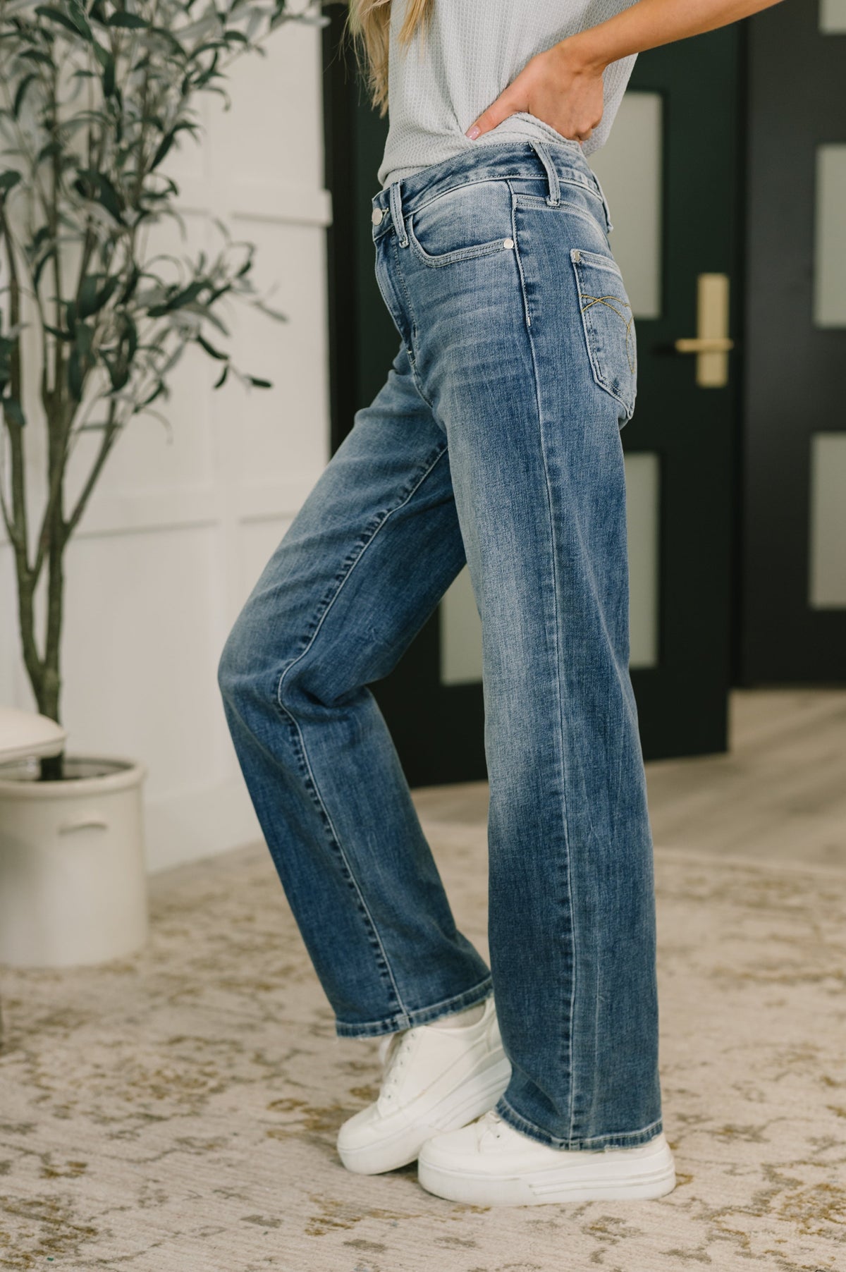 Person wearing blue jeans and white sneakers indoors with a plant and door in the background