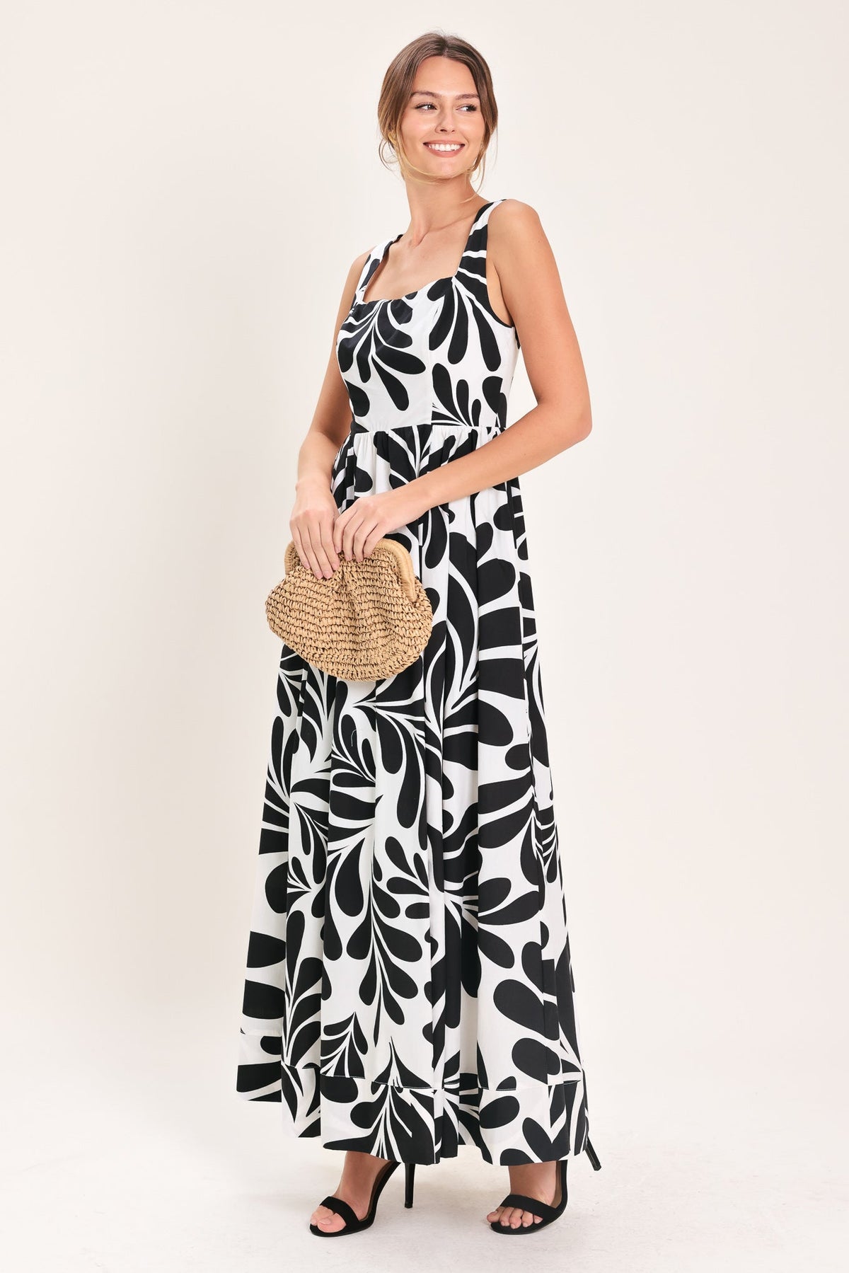 Woman wearing a black and white patterned dress holding a straw bag on a white background