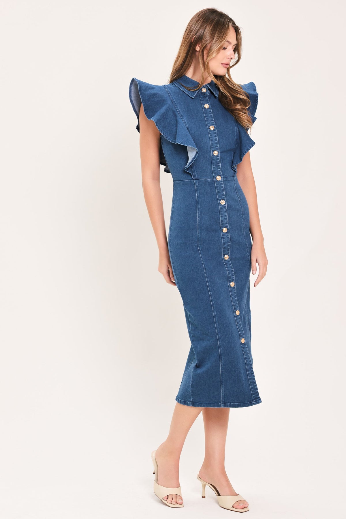 A woman modeling a blue denim midi dress with ruffled cap sleeves, a button-down front, and a fitted silhouette, paired with beige heeled sandals against a plain light background.