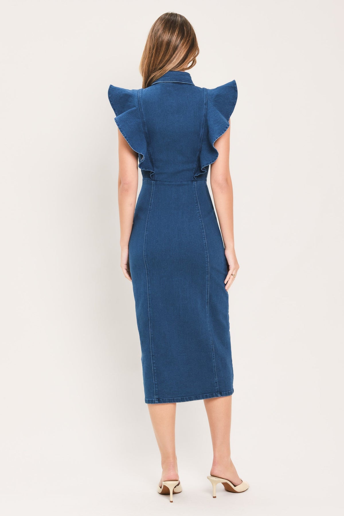 A woman viewed from the back wearing a blue denim midi dress with dramatic ruffled shoulder details and a tie at the waist, paired with beige heeled sandals against a plain light background.