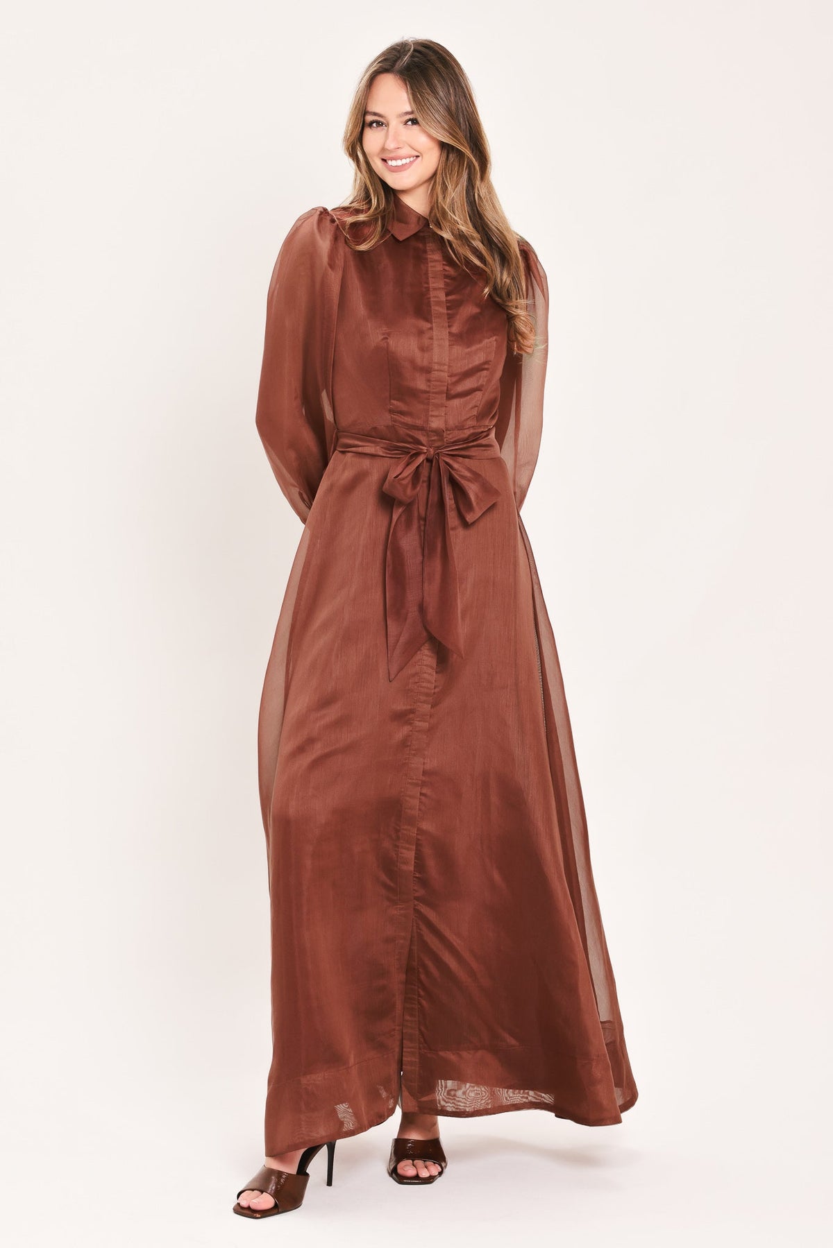 A woman modeling a long-sleeved, rust-brown maxi shirt dress with a matching waist tie, photographed against a solid off-white studio background.