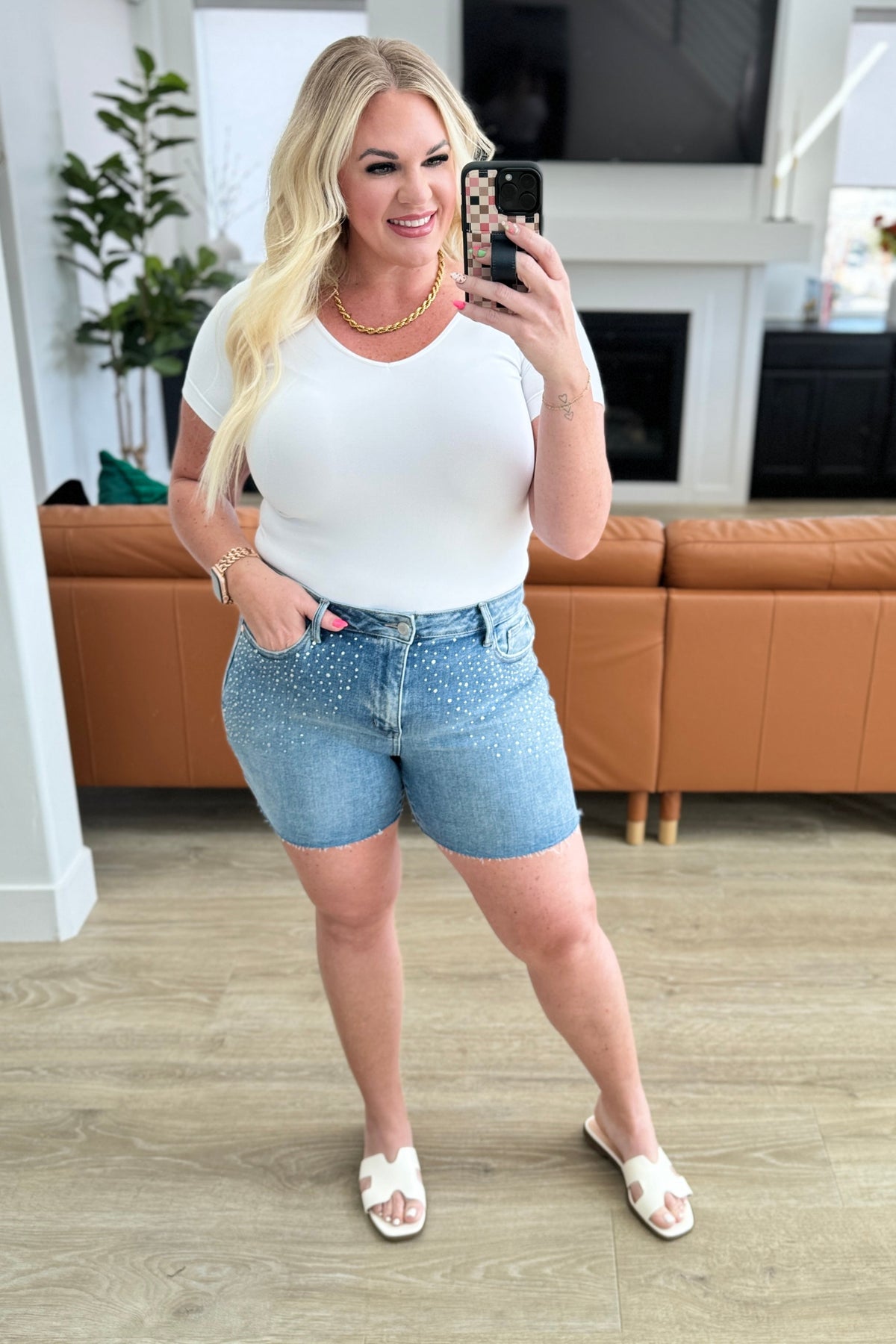 Woman taking a selfie in a living room wearing a white top and denim shorts.