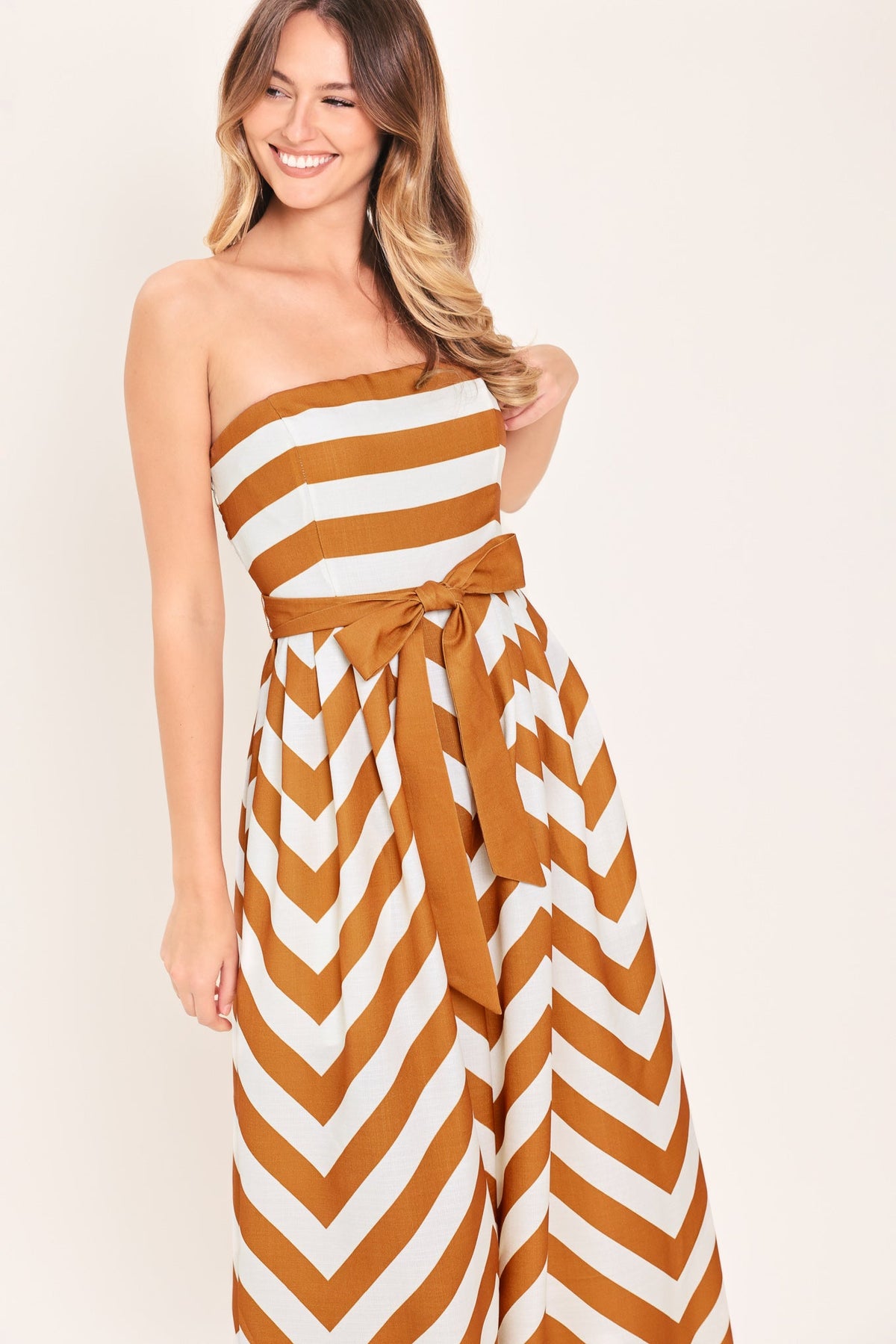 Woman wearing a strapless, brown and white striped jumpsuit against a plain background