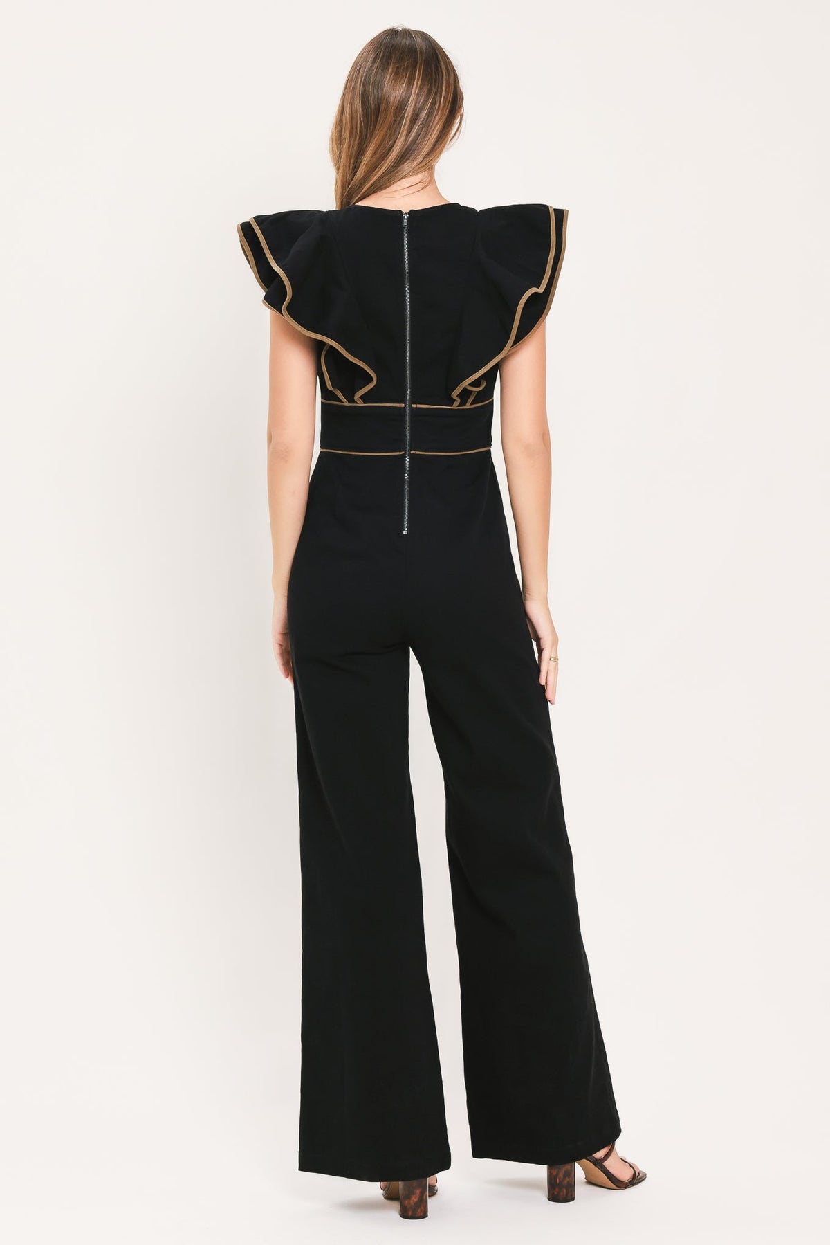 A woman shown from the back wearing a black wide-leg jumpsuit with dramatic ruffled shoulder details and gold contrast trim, photographed against a plain light background.