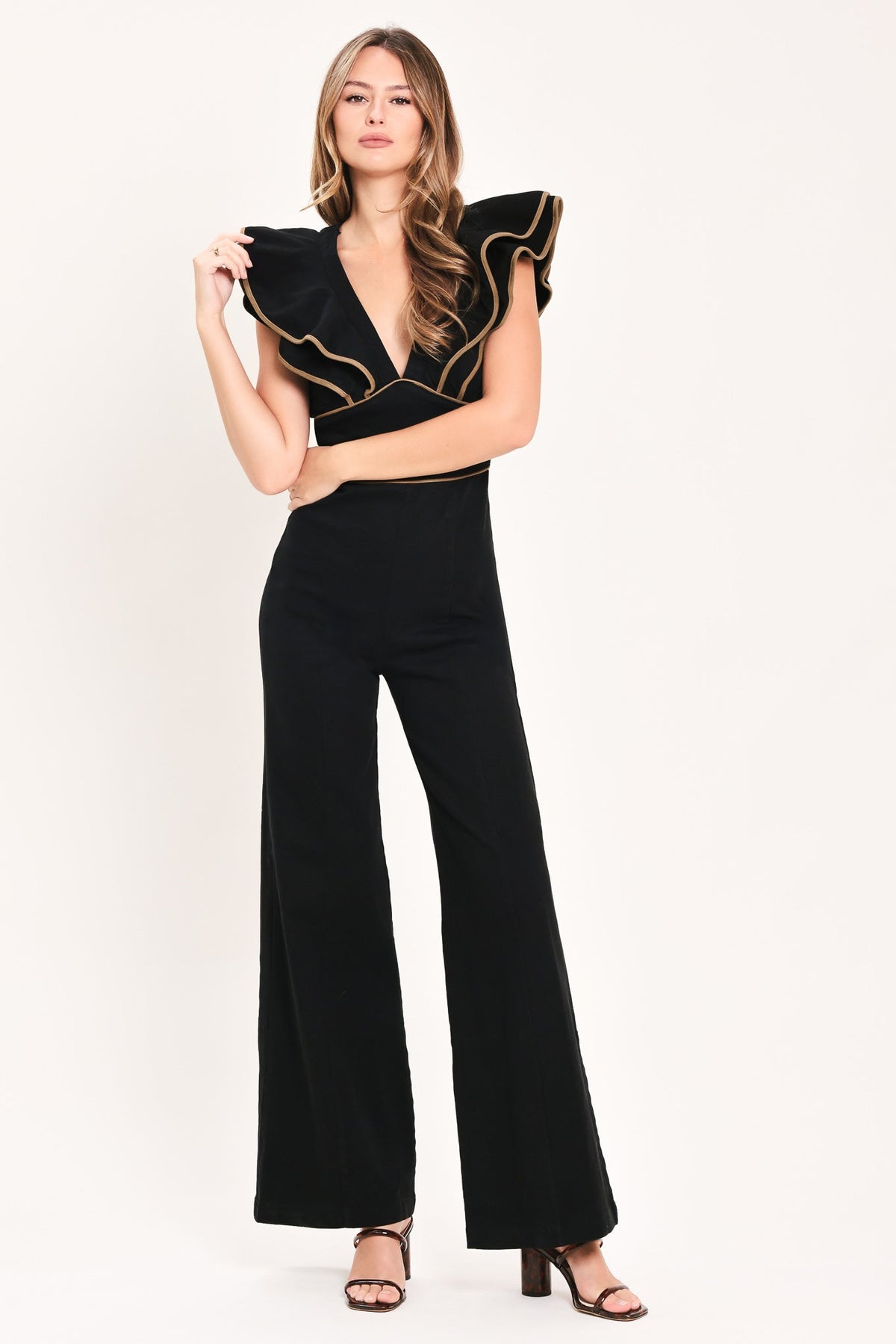 A woman modeling a black wide-leg jumpsuit with dramatic ruffled shoulder details and gold trim, paired with brown heeled sandals, against a plain off-white studio background.