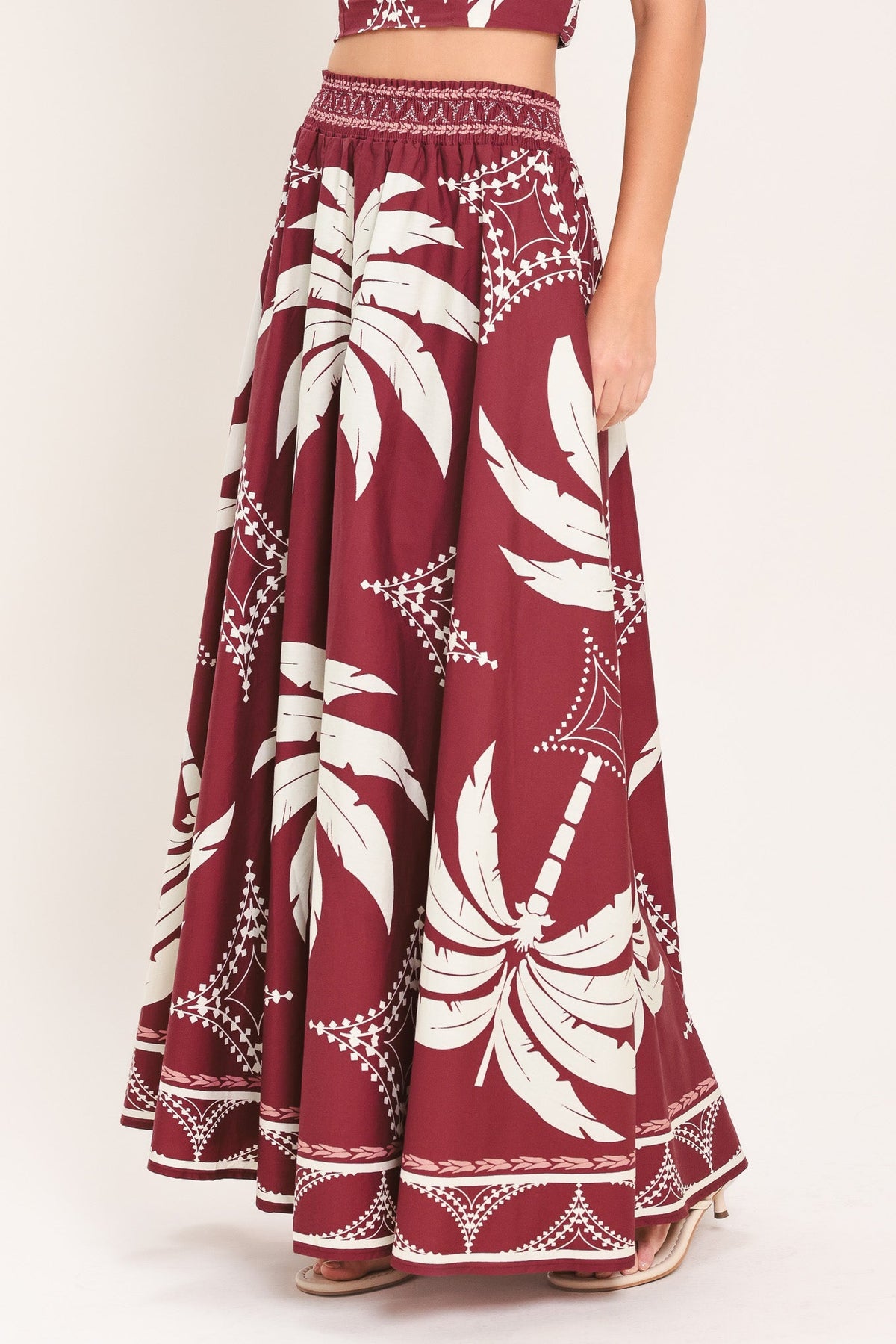 A maroon maxi skirt with a white palm tree and geometric tribal pattern, shown on a model against a light beige background.