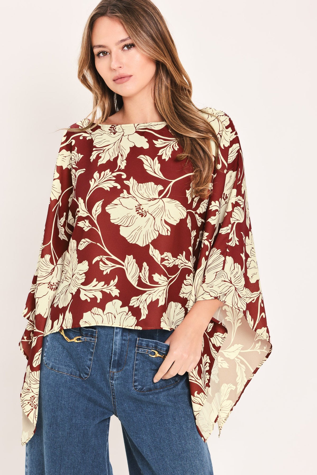 A woman wearing a burgundy and cream floral print blouse with wide bell sleeves, paired with blue jeans, against a plain light background.