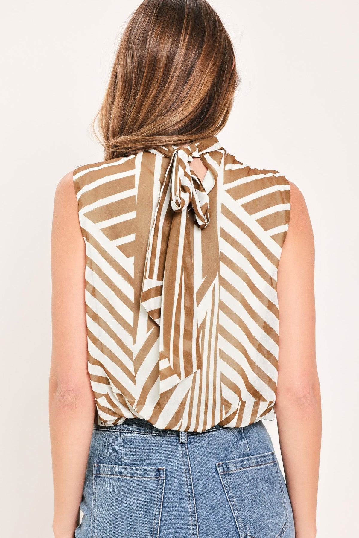 A woman shown from the back wearing a sleeveless top with a brown and cream geometric pattern and a tie-neck detail, paired with blue jeans against a light beige background.