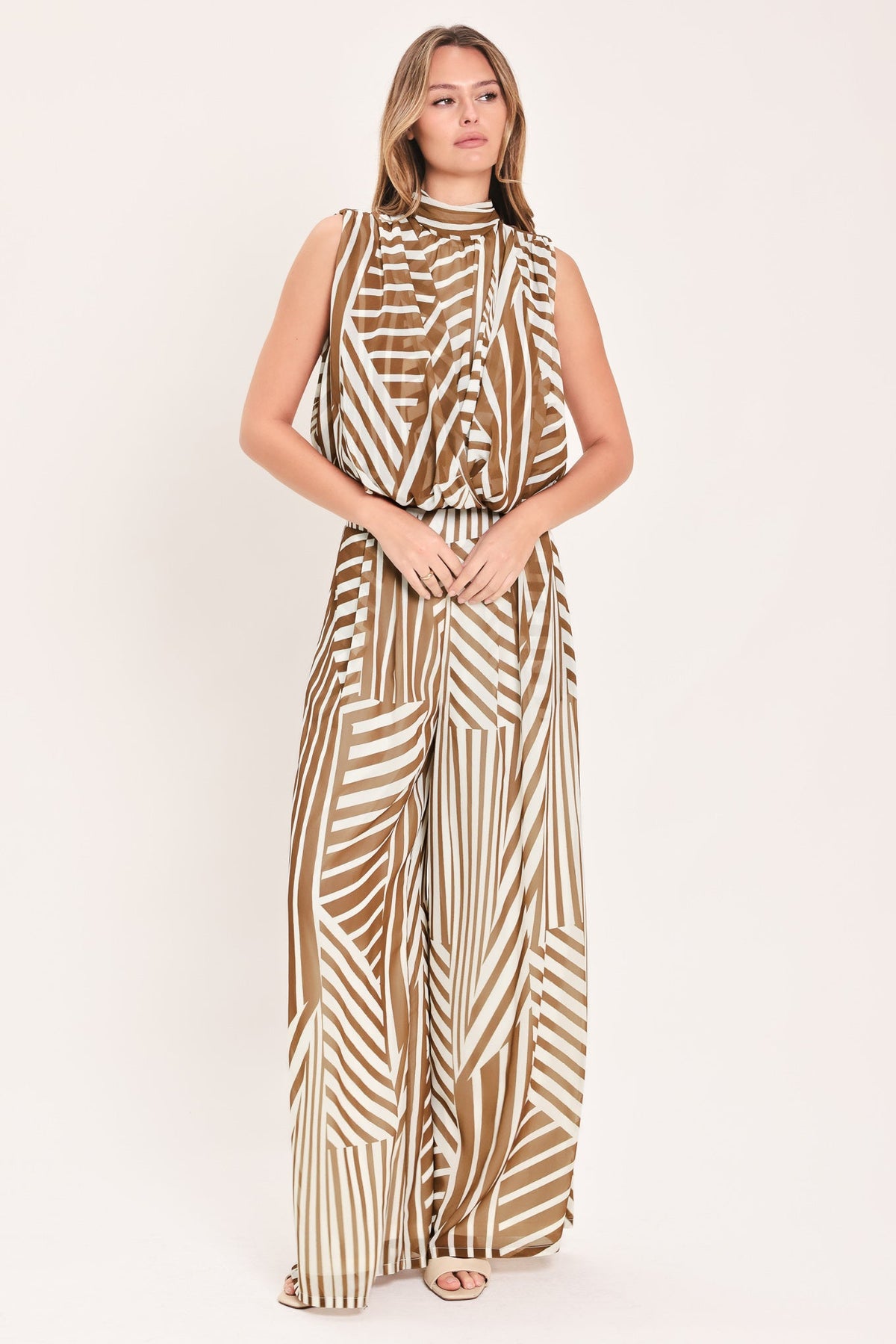 A woman modeling a sleeveless high-neck jumpsuit with a brown and cream abstract geometric pattern, standing against a plain light beige studio background.