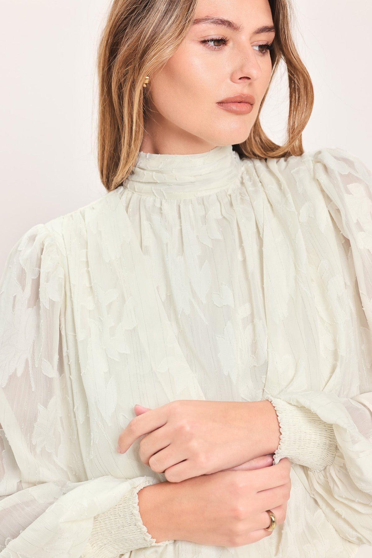 A close-up of a woman wearing a high-neck, long-sleeve blouse in a light sage green or off-white color with a textured, slightly sheer overlay and gathered details.