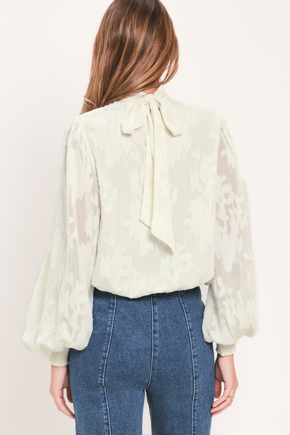 A woman viewed from the back wearing a cream-colored lace blouse with a large bow at the back of the neck and blue jeans, set against a plain off-white background.