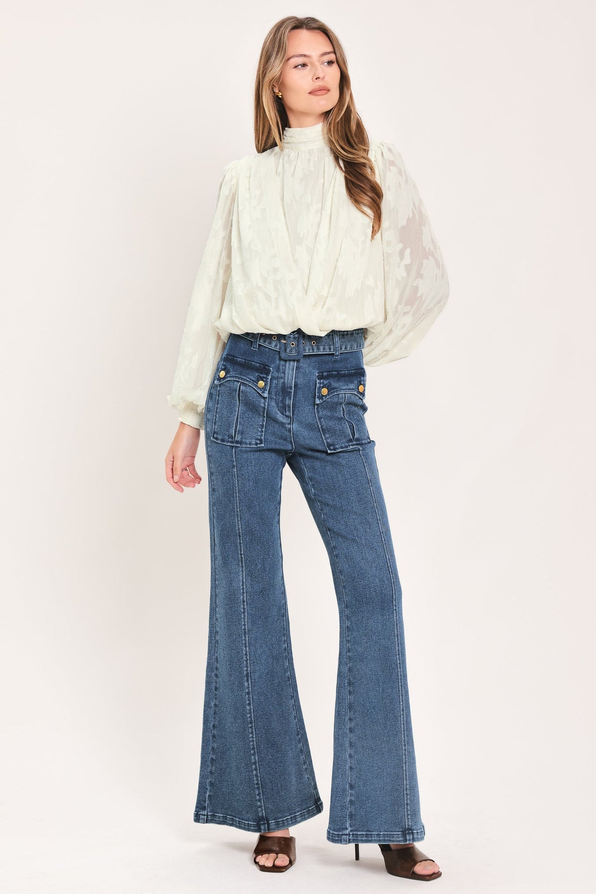 A woman modeling a cream-colored, long-sleeved lace blouse paired with high-waisted, wide-leg blue denim jeans and dark brown heeled sandals, set against a plain off-white studio background.
