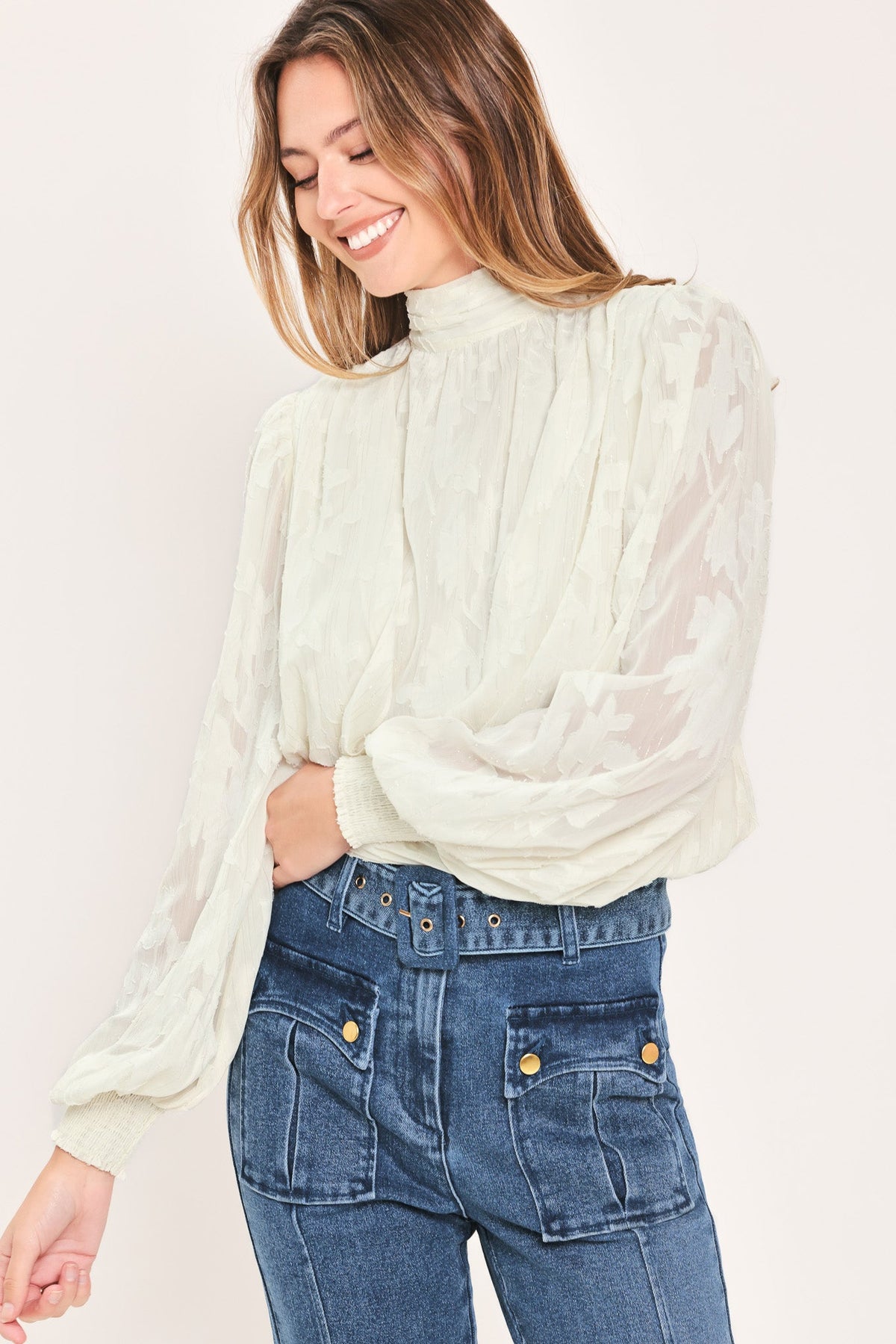 A woman wearing a cream-colored, long-sleeved sheer blouse with a high gathered neckline and blue denim jeans, photographed against a solid off-white background.