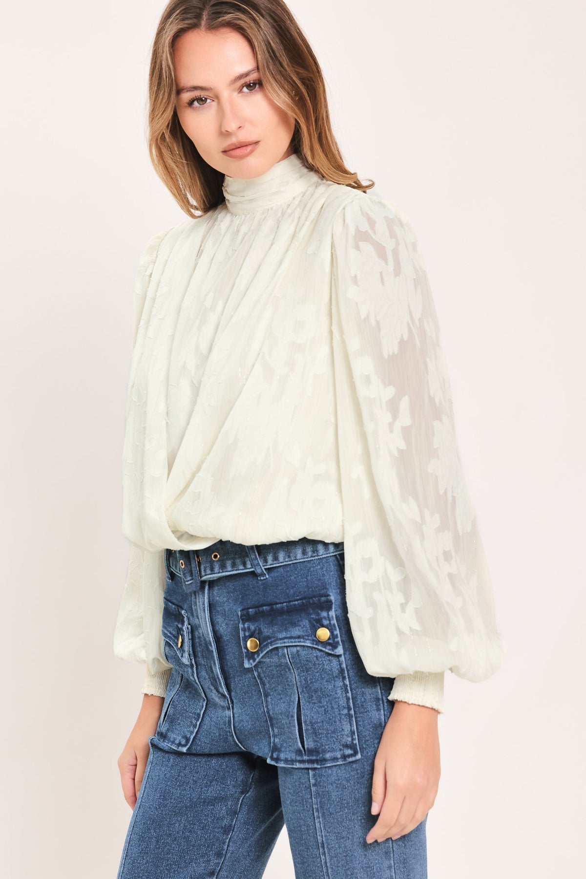 A woman wearing a cream-colored, sheer lace long-sleeve blouse with a high mock neck and blue jeans, photographed against a solid light beige background.