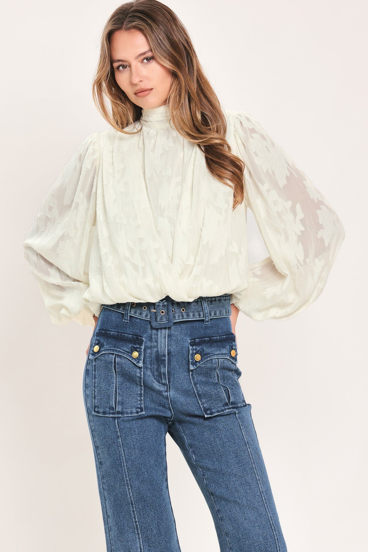 A woman wearing a cream-colored, semi-sheer long-sleeve blouse with a high neck and blue jeans, photographed against a solid light beige background.