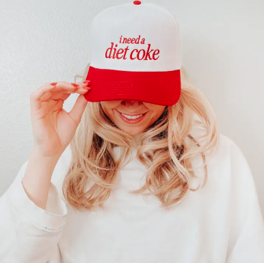A woman wearing a white and red trucker hat with the text 'i need a diet coke' in red script on the front panel, holding the brim over her eyes. She is wearing a white sweatshirt against a plain light background.