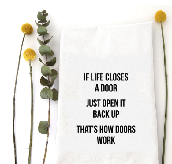A white kitchen towel with black printed text reading 'IF LIFE CLOSES A DOOR JUST OPEN IT BACK UP THAT'S HOW DOORS WORK', styled flat-lay with eucalyptus stems and yellow flowers on a white background.