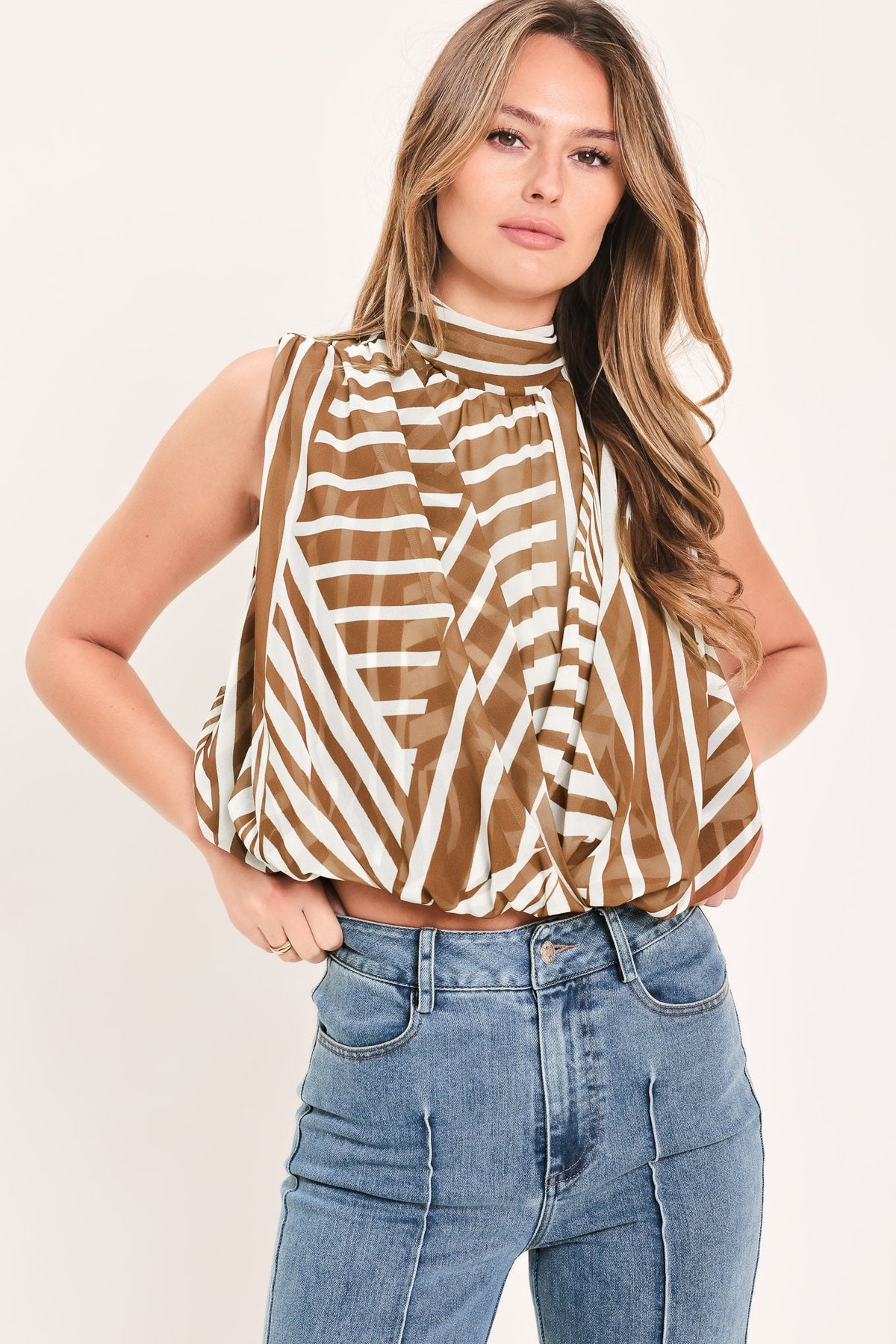 A woman modeling a sleeveless mock-neck top with a brown and white diagonal striped pattern, paired with blue jeans, against a plain light background.